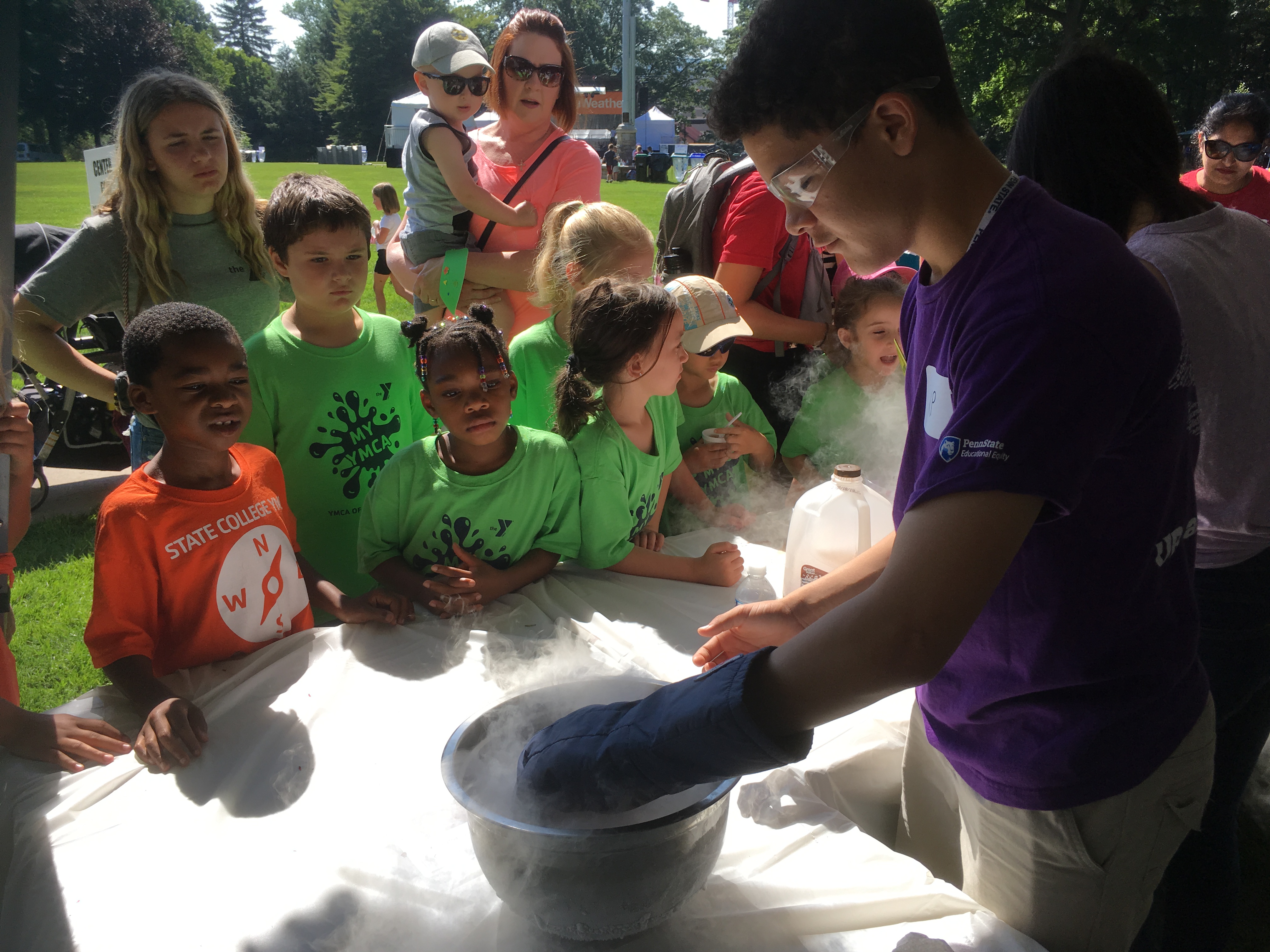 Children gather around a scientific demonstration on a table outdoors.